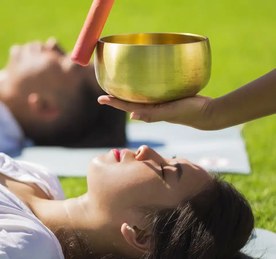 A person holds a singing bowl above a woman lying on the grass with her eyes closed, while another person lies nearby, both appearing to participate in a sound healing or meditation session outdoors. A person holds a singing bowl above a woman lying on the grass with her eyes closed, while another person lies nearby, both appearing to participate in a sound healing or meditation session outdoors.