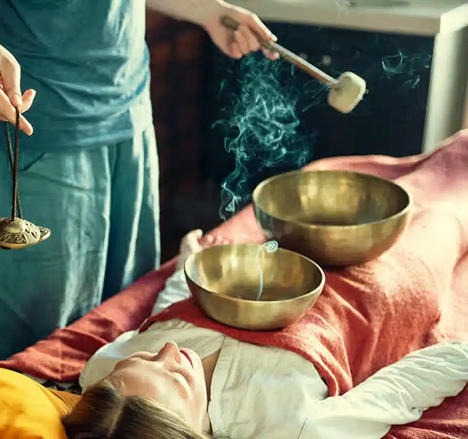 A person lies on a table covered with an orange blanket, with metal singing bowls placed on their body. Another person is striking a bowl and holding incense, creating smoke, suggesting a sound healing or relaxation session. A person lies on a table covered with an orange blanket, with metal singing bowls placed on their body. Another person is striking a bowl and holding incense, creating smoke, suggesting a sound healing or relaxation session.